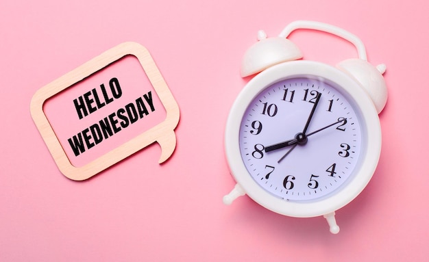 Op een delicate roze achtergrond, een witte wekker en een houten frame met de tekst HELLO WEDNESDAY