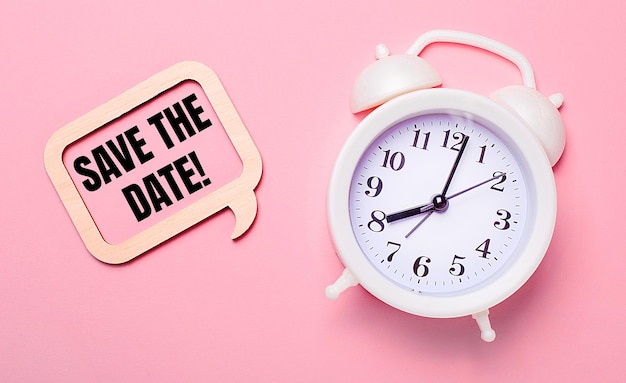 Foto op een delicate roze achtergrond een witte wekker en een houten frame met de tekst save the date