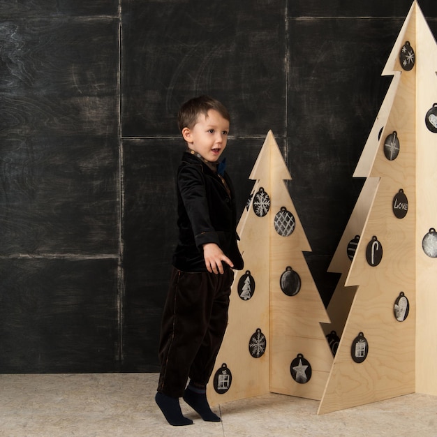 Op een donkere achtergrond poseert kleine jongen in een stijlvol kostuum in de buurt van een kerstboom gemaakt van hout