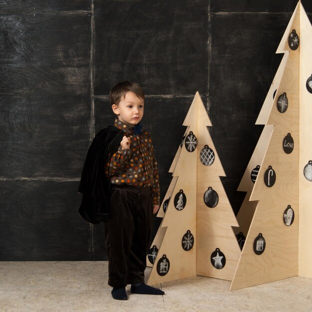 Op een donkere achtergrond poseert kleine jongen in een stijlvol kostuum in de buurt van een kerstboom gemaakt van hout