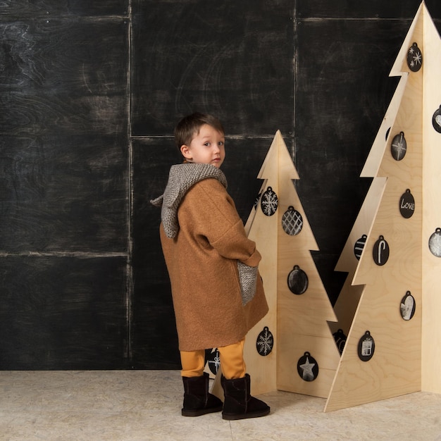 Op een donkere achtergrond poseert kleine jongen in een stijlvolle jas bij een kerstboom van hout