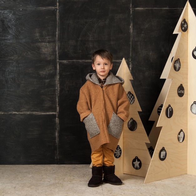 Op een donkere achtergrond poseert kleine jongen in een stijlvolle jas bij een kerstboom van hout