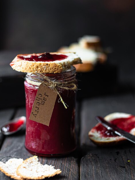 Op een houten tafel staat een potje kersenjam met brood en een lepel jam