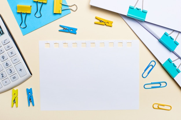 Op een licht oppervlak bevindt zich een rekenmachine, gele en blauwe paperclips, wasknijpers, documenten en een blanco vel papier om tekst in te voegen. Plat leggen. Plaats om tekst in te voegen.