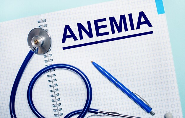 Op een lichtblauwe achtergrond, een open notitieboekje met het woord ANEMIA, een blauwe pen en een stethoscoop