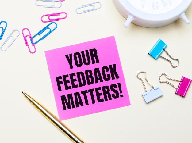 Op een lichte achtergrond een witte wekker, roze, blauwe en witte paperclips, een gouden pen en een roze sticker met de tekst YOUR FEEDBACK MATTERS