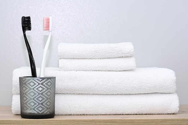 Op een plank in de badkamer liggen witte handdoeken van verschillende formaten netjes opgevouwen en een glazen beker met zwarte en roze tandenborstels. Het concept van ochtendprocedures, bestel thuis, familie.