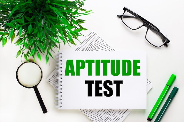 Op een witte achtergrond ligt een notitieboekje met het woord APTITUDE TEST, bril, vergrootglas, groene markeringen en een groene plant