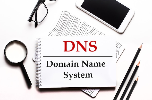 Op een witte achtergrondbril, een vergrootglas, potloden, een smartphone en een notitieboekje met de tekst DNS Domain Name System