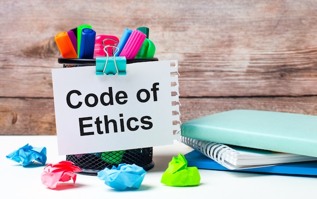 Op het bureaublad en tegen de achtergrond van een houten muur staat een standaard met veelkleurige viltstiften, heldere stukjes papier en een vel papier met de tekst code of ethics