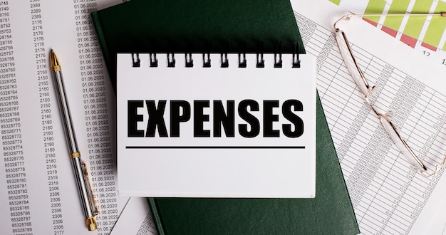 Op het bureaublad staan rapporten, een bril, een pen, een groen dagboek en een wit notitieboekje met de woorden EXPENSES. Werkplek close-up. Bedrijfsconcept