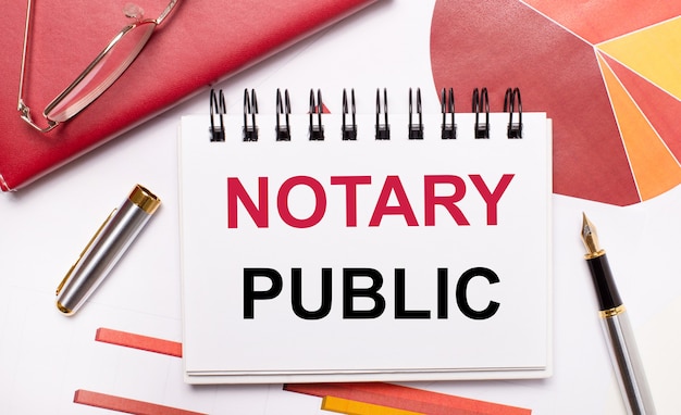 Op het bureaublad staat een wit notitieboekje met de tekst NOTARY PUBLIC, een pen, bordeauxrode en rode tafels en een bril met gouden montuur. Bedrijfsconcept.