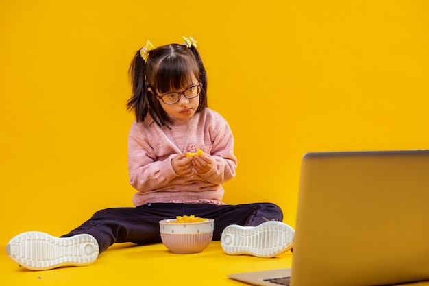 Op kale vloer. Geïnteresseerde kleine dame met het syndroom van Down chips eten en tekenfilms kijken op laptop