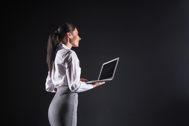 Op kantoor. Vrolijke gelukkige positieve zakenvrouw die een laptop vasthoudt en naar het grote computerscherm kijkt terwijl hij glimlacht