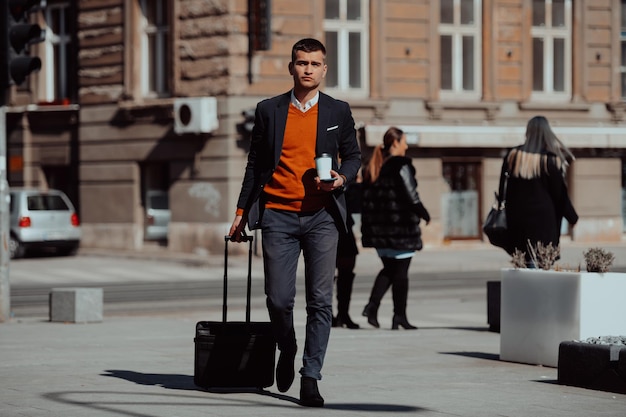 Op weg naar de luchthaventerminal. Jonge zelfverzekerde zakenmanreiziger die door de straten van de stad loopt en zijn koffer trekt die koffie drinkt en op een smartphone spreekt. Hoge kwaliteit foto