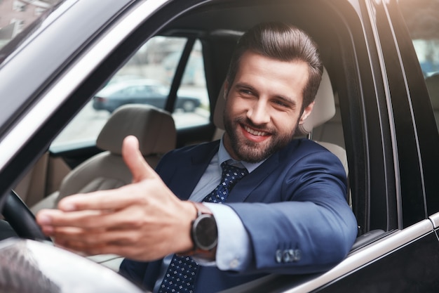 Op weg naar zakelijke bijeenkomst. Gelukkig en knappe jonge zakenman in volledig pak is gebaren en glimlachen terwijl hij in zijn auto zit. Positieve emotie. Bedrijfsconcept.