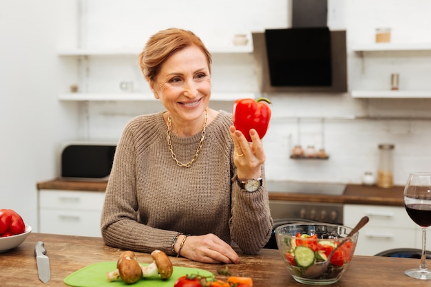 Op zoek op peper. Stralende rijpe dame die op houten tafel in de keuken leunt en hele peper presenteert