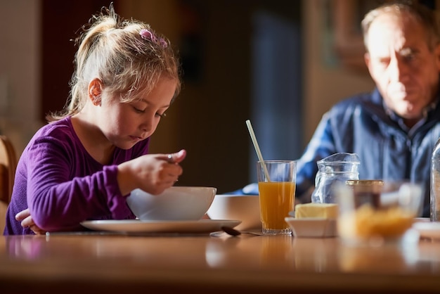 Opa's houden de ontbijttijd in de gaten Bijgesneden opname van een jong meisje dat ontbijtgranen eet aan tafel met haar grootvader op de achtergrond