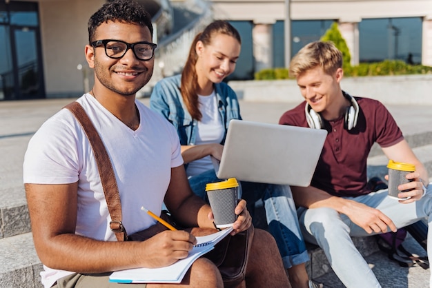 Open-air lecture. Two friends sitting on the background and keeping smile on faces while looking at laptop