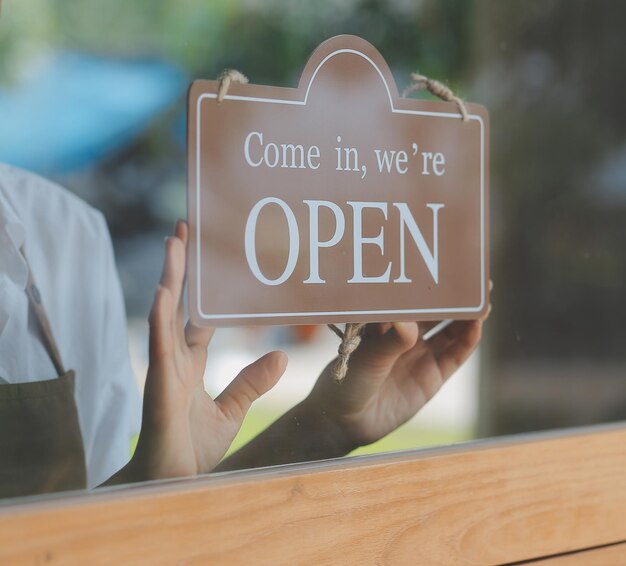 Foto aprire il barista cameriera donna che indossa la maschera facciale di protezione girando il cartello aperto sulla porta di vetro nel moderno caffè caffetteria caffetteria ristorante negozio al dettaglio piccolo imprenditore cibo e bevande concetto