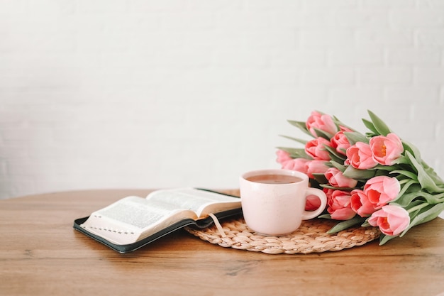 Open bible with tulip flowers and a cup of tea on light wood