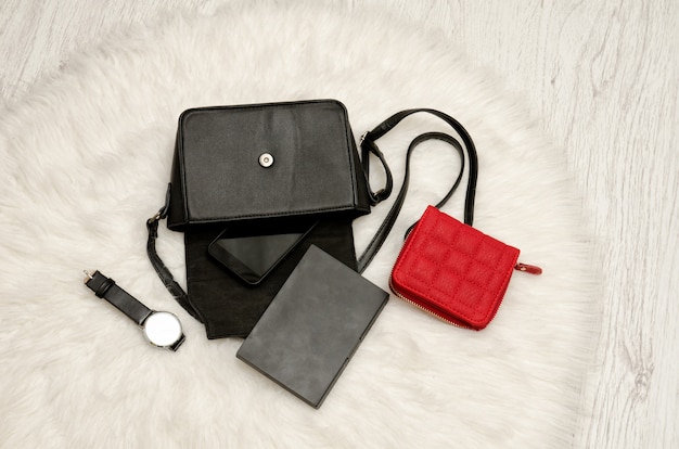 Открытая черная сумка с брошенными вещами, блокнотом, мобильным телефоном, часами и красной сумочкой.