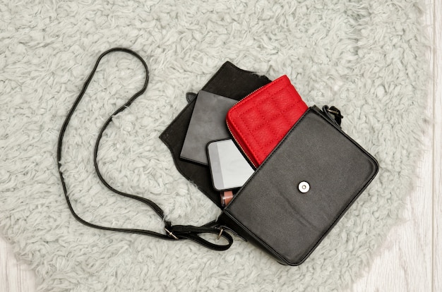 Open black handbag, red purse, mobile phone and lipstick in it. Grey fur background, top view