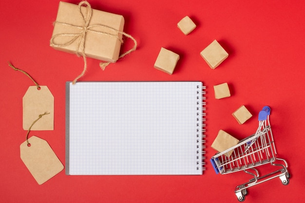 Open blank notebook with place to add text, shopping cart and boxes on a bright red background. Online shopping concept