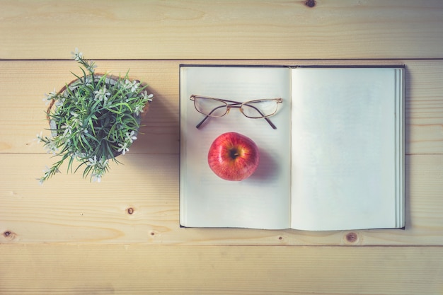 Open book, apple and glasses on wooden table with flower vase