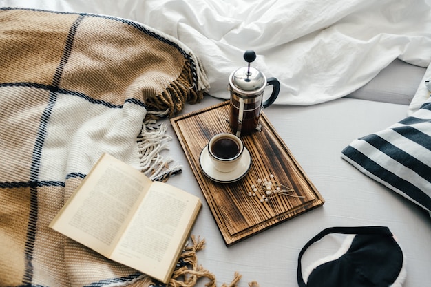Open book in bed with coffee brewed in a french press and cup on a wooden board