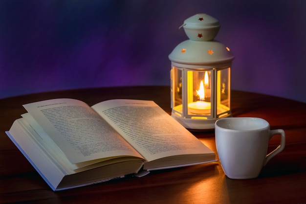an open book and a cup of tea or coffee at the light of a lantern with a candle in the evening
