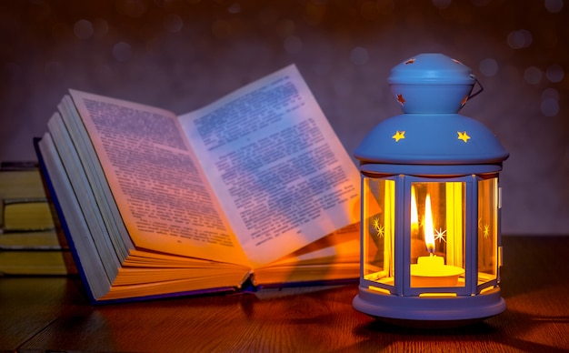 提灯の明かりで照らされた開いた本。開いた本の近くのキャンドルとランタン。ろうそくの明かりで読書。ロマンチックな夜