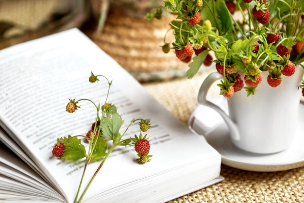 Open book laying near summer bunch of wild strawberry twigs placed in tea mug