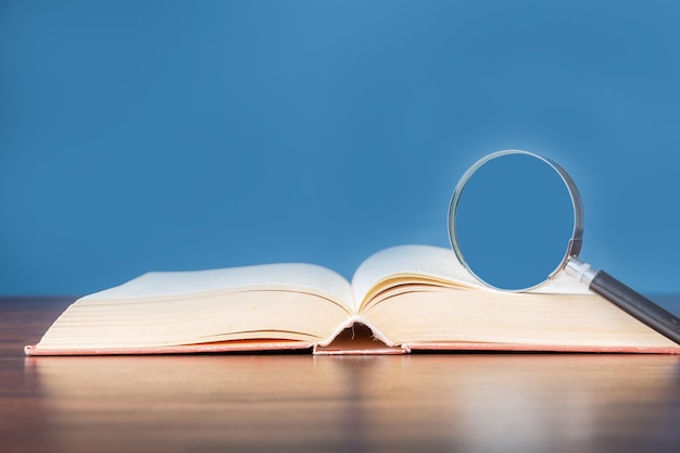 Open book with magnifying glass on wooden desk in information library of school or university concept for educationreading study copy space and blue background