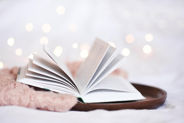 Open book on wooden tray with knitted sweater closeup over Christmas lights