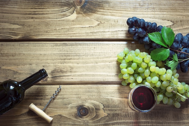 Open bottle of red wine with wineglass, corkscrew and ripe grape on wooden board.