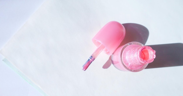 An open bottle with pink nail polish and a brush next to it on a white surface with a shadow the concept of fashionable beauty