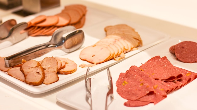 Open buffet at the hotel. Variety of ham and sausage on white plates with tongs