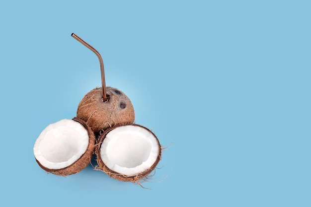 Open coconut with straw on blue background. Tropical drink, summer vacation minimal creative concept. Cracked coconut fruit with coconut milk inside, copy space for text.