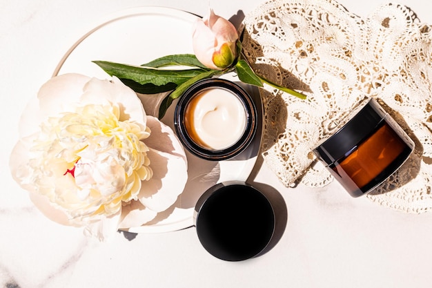 An open dark glass jar with a cosmetic selfcare product on a marble white background and a peony bud natural cosmetics