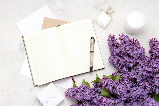 Open diary, pen, gift box, perfume bottle, Candle, envelopes and a twig of lilac on a white stone surface. Flat lay, top view