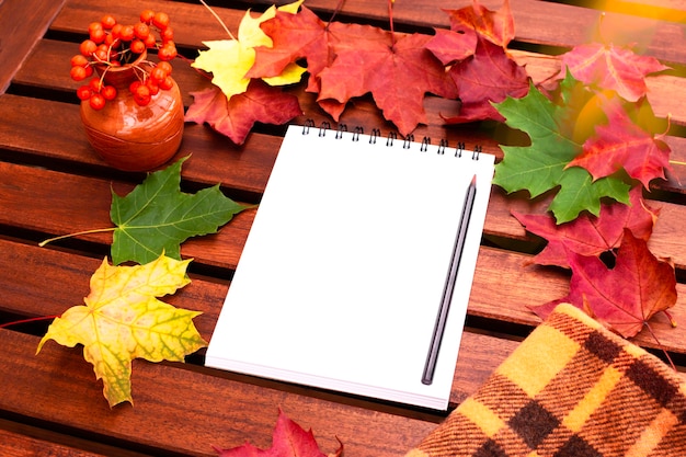 Open een leeg notitieboekje met een potlood op een houten tafel met herfst esdoorn bladeren verspreid en een vaas met lijsterbes.