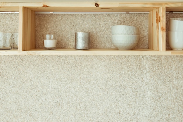 Открытая плавающая деревянная полка с керамическими тарелками и стаканами, висящая на белой стене в современной дизайнерской кухне. Экологичный уютный минималистский дом с концепцией