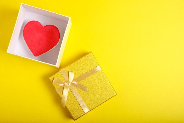 Open gift box in gold color with ribbon and bow with heart inside on yellow background