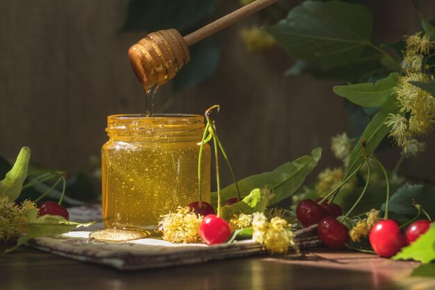 Open glass jar of liquid honey and honey dipper bunch of linden flowers and red cherry on wooden surface Ray of sunlight Dark rustic style