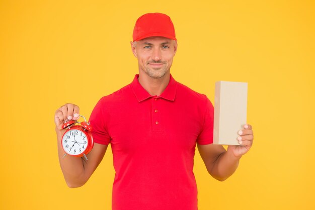 Open hours Time is value Delivered to your destination Service delivery Courier and delivery Postman delivery worker Handsome man red cap hold alarm clock yellow background Delivering purchase