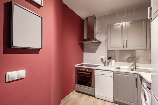 Open kitchen with white appliances dishwasher washing machine and oven and stainless steel extractor hood with white stone countertop red painted wall and one with gray tiles