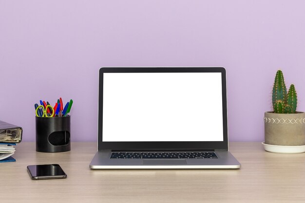 Open laptop with smartphone, cactus and business folders on a wooden table against a lilac wall, office work concept