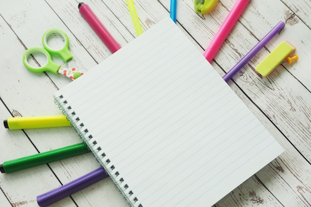 An open notebook, colorful bright markers, pens, sharpener, eraser, scissors, stickers and clay on a wooden background.