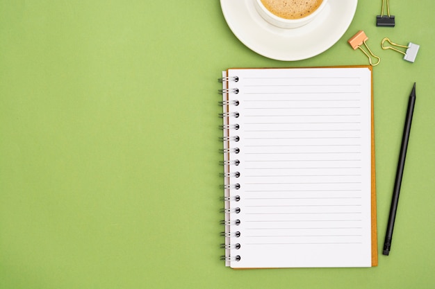 Open notebook with empty page and coffee cup. Table top, work space on green background. Creative flat lay.